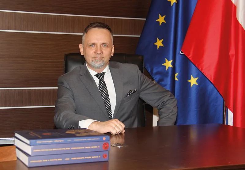 Mężczyzna w garniturze siedzi za biurkiem obok flagi Unii Europejskiej i Polski, przed nim leżą trzy książki.